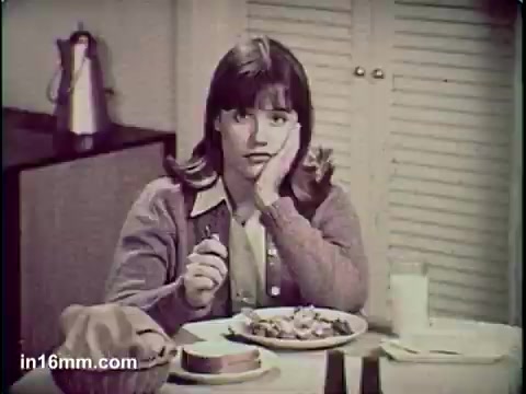 The still from the 'Total Cereal Commercial' features a young girl sitting at a dining table, looking bored or disinterested as she rests her chin on her hand. She has a plate of cereal in front of her, alongside a glass of milk and what appears to be a slice of bread. The setting conveys a typical breakfast scene, but her expression suggests dissatisfaction or a lack of enthusiasm about the meal. The overall tone is reminiscent of mid-20th century advertising.