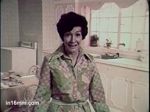 The still from the 'Total Cereal Commercial' features a woman in a colorful, patterned dress with a thoughtful expression, set in a bright, retro kitchen. The scene conveys a warm, homey atmosphere typical of vintage commercials, likely promoting a healthy breakfast option.