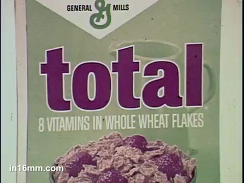 The still features a box of Total cereal, prominently displaying the brand name 'total' in bold purple letters. Below the name, it highlights '8 vitamins in whole wheat flakes,' emphasizing its nutritional value. The background is a soft green, providing a fresh aesthetic that complements the cereal's health-focused message. The cereals appear textured and are garnished with fruits, likely to appeal to a health-conscious audience.
