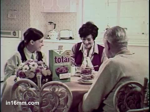 The still from the 'Total Cereal Commercial' features a family scene set around a dining table. A young girl and a woman, likely a mother or relative, share a moment with an older man, possibly a grandfather. They are engaged in conversation, with a box of Total cereal prominently displayed on the table. The environment appears warm and inviting, reflecting a typical breakfast setting, with subtle decor that suggests a mid-20th century aesthetic.