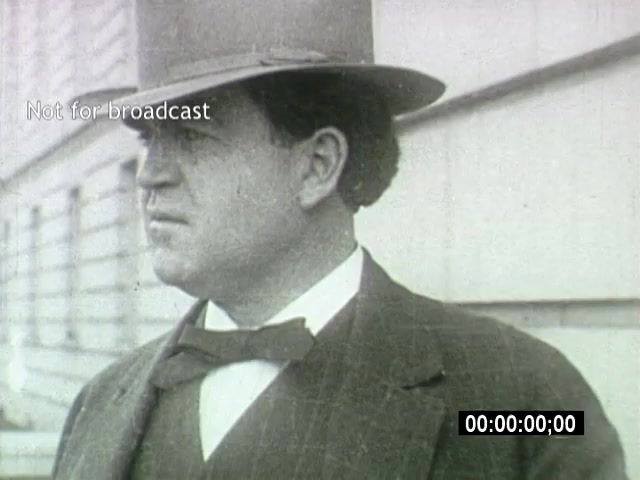 The still from 'John L Lewis 1919 Miner Strike footage' features a man in formal attire, wearing a bow tie and a fedora hat. He appears to be outdoors, possibly addressing the situation surrounding the miner strike. The setting conveys a sense of seriousness and formality associated with labor movements of that era. The footage is not for broadcast, and there is a timestamp indicating the timing of the clip.