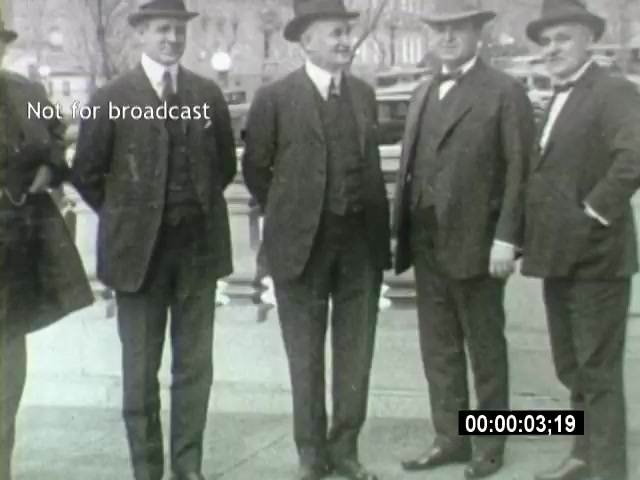 The still from the 'John L Lewis 1919 Miner Strike footage' depicts a group of four well-dressed men, all wearing suits and hats, standing together in an urban setting. They appear to be engaged in conversation. The backdrop suggests a busy public area, possibly during the time of the miner strike, reflecting the socio-political climate of the era. The image is black and white, typical of the early 20th century footage.