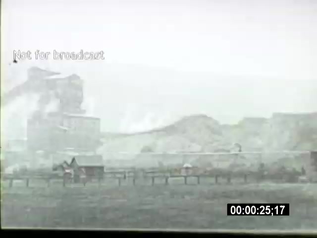 The still from the 'John L Lewis 1919 Miner Strike footage' captures a scene depicting industrial facilities, likely a coal mine or related infrastructure, enveloped in a misty atmosphere. In the foreground, there's a wooden structure, possibly a small shed, beside a fence. The background features smoke or steam emanating from the industrial complex, indicating active operations despite the strike. The monochrome tones convey a sense of historical significance and the gritty nature of mining life during that era. The text 'Not for broadcast' suggests the footage is intended for archival or educational use rather than public airing.