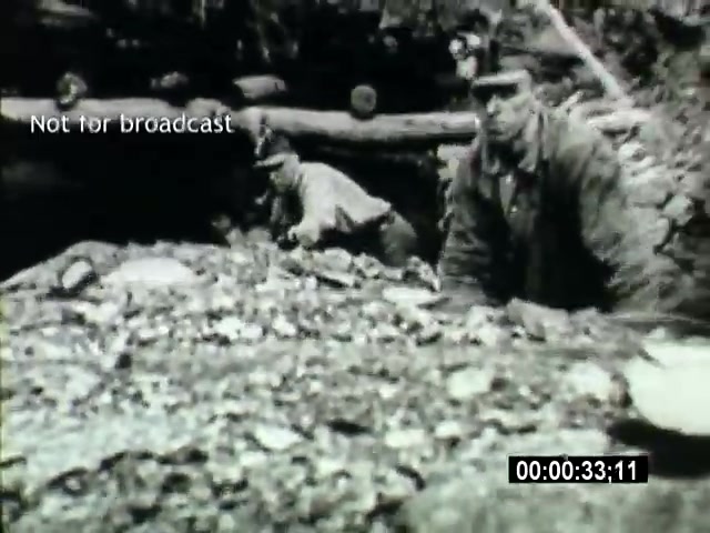 The still shows two miners engaged in manual labor during the 1919 miner strike. They appear focused on their work, with one miner crouched down and the other visible behind him, suggesting a challenging environment. Both are wearing traditional miners' attire and helmets, reflecting the historical context of mining operations during this period. The image captures the gritty reality of their work and the economic struggles faced by miners at the time. A watermark reading 'Not for broadcast' is present, along with a time code.