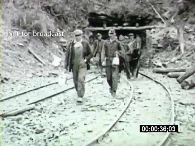 The still from the 'John L Lewis 1919 Miner Strike footage' depicts a group of miners walking along railway tracks, emerging from a tunnel. They are dressed in work attire typical for the time, likely indicating the era's mining conditions. The scene reflects the labor struggles and solidarity of miners during the 1919 strike, capturing a moment of their daily lives and the broader labor movement. The stark black-and-white imagery highlights the historical context of early 20th-century coal mining.