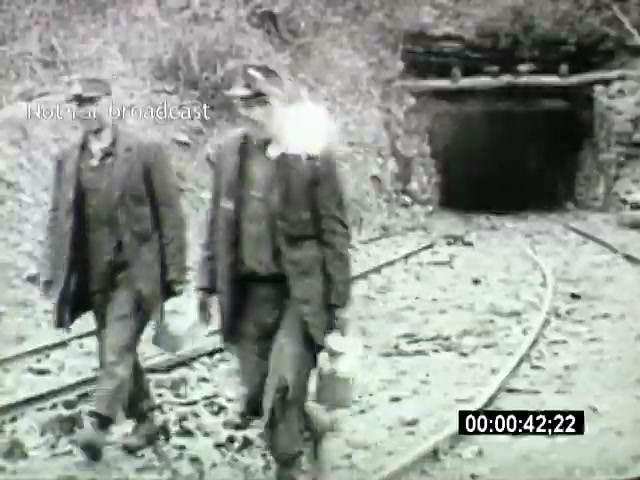 The still from the 'John L Lewis 1919 Miner Strike footage' depicts two miners walking along railway tracks near a mine entrance. They are dressed in typical early 20th-century work attire, including hats and heavy coats, and each carries a lunch pail. The setting is gritty and industrial, emphasizing the harsh conditions faced by miners during this period of labor unrest. The image captures the resilience and camaraderie of the mining community during the strike.