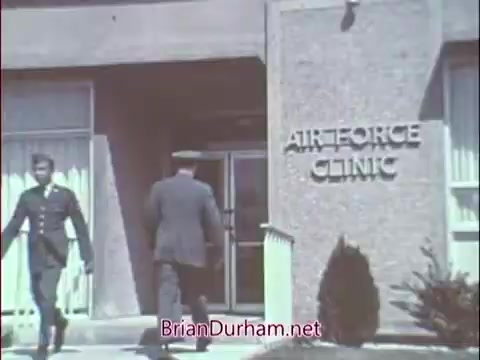 The still features an exterior shot of an Air Force clinic, showcasing a modern building with large windows. Two military personnel, one in a uniform and another in a suit, are exiting the entrance. The signage prominently displays 'AIR FORCE CLINIC,' indicating the military context of the scene, likely aimed at promoting medical careers within the Air Force during the Vietnam era.