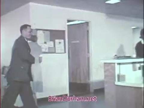 The still appears to show a scene from a 16mm commercial aimed at recruiting doctors for the Air Force Reserve during the Vietnam era. It features a man in military attire walking through a modern, professional-looking office environment. There are individuals in the background, suggesting a healthcare or administrative setting. The overall tone is formal and focused on military professionalism and collaboration.