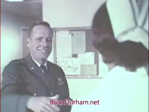 The still depicts a male Air Force officer in uniform smiling and extending his hand for a handshake to a nurse, who is dressed in traditional medical attire. The background suggests a hospital or medical facility, indicative of the collaborative environment between military and medical personnel during the Vietnam era. This scene likely emphasizes camaraderie and recruitment for military doctors.