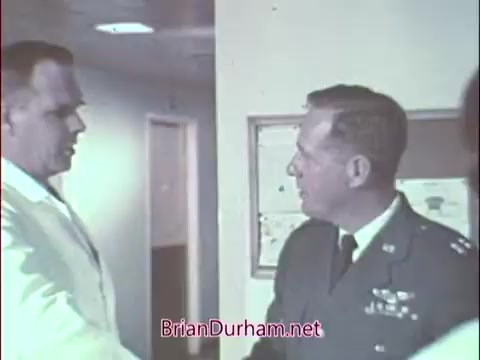 The still features two men engaged in a handshake in an interior setting, likely a medical facility. One man is dressed in a white lab coat, indicating a medical professional, while the other is wearing a military uniform, suggesting he is associated with the Air Force Reserve. The environment appears clinical, reflecting a recruitment theme aimed at attracting doctors to military service. The overall tone is professional and cordial.