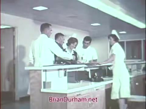 The still features a group of healthcare professionals, including doctors and a nurse, gathered around a reception desk in a hospital or medical setting. They appear engaged in discussion or collaboration, highlighting teamwork and communication. The scene reflects the medical environment of the Vietnam era, likely aiming to illustrate the role of doctors in the Air Force Reserve within the context of military service. The color palette is characteristic of mid-20th century film, giving it a vintage feel.