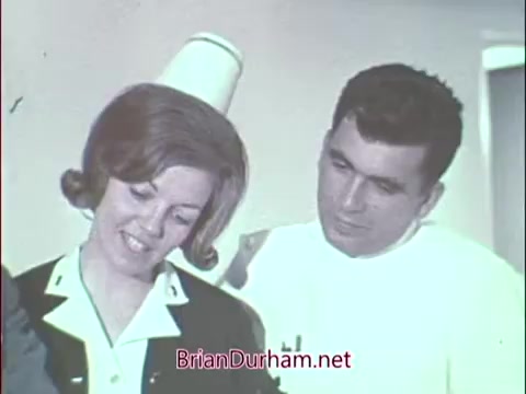 The still features a nurse and a doctor engaging in a professional setting, likely a hospital or medical facility. The nurse, wearing a traditional uniform with a white collar and a cap, is looking down at a document or an object in her hands, while the doctor leans in, appearing attentive. The scene conveys a collaborative atmosphere typical of medical environments during the Vietnam era, emphasizing the role of healthcare professionals in the Air Force Reserve.