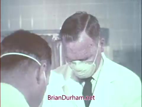 The still depicts two medical professionals in a clinical setting, both wearing surgical attire, including masks and gloves. One figure is focused on a task in front of him, while the other appears to be observing or assisting. The environment suggests a sterile medical atmosphere, likely during an operation or medical procedure, indicative of the Air Force Reserve's focus on recruiting doctors during the Vietnam era.