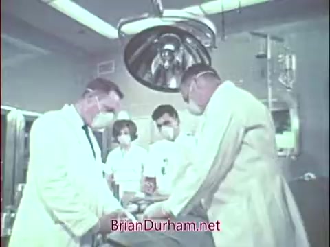 The still from the 'Vietnam era Air Force Reserve recruiting commercial for doctors' depicts a medical scene in a surgical room. It shows several medical professionals, dressed in white coats and masks, focused on a patient laying on an operating table. The environment suggests a serious and professional atmosphere, reflecting the high stakes of medical work during the era. The image is likely intended to highlight the role of doctors in the Air Force Reserve, emphasizing their contributions to both military and civilian healthcare.