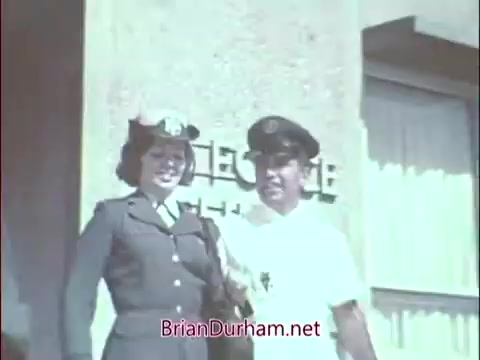 The still features two individuals in military attire standing outside a building. The woman, dressed in a uniform with a hat, and the man, in a white uniform, appear to be engaged in a friendly conversation or photo opportunity. The background includes a sign that likely indicates a medical or recruitment facility, aligning with the theme of the Air Force Reserve recruiting commercial for doctors from the Vietnam era. The overall setting reflects a professional yet welcoming atmosphere.