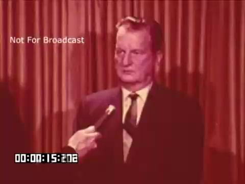 The still shows a middle-aged man in a suit, standing in front of a textured curtain, seemingly in a press interview. A microphone is directed towards him, indicating he is answering questions, likely about the topic of auto-color in film or broadcast. The muted color tones suggest it may be an older footage, possibly highlighting a discussion on the preferences of color correction technology in media.