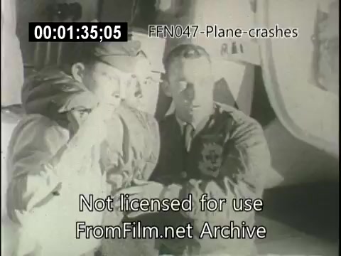The still features two men, dressed in military attire, closely examining something on a surface, possibly discussing the aftermath of a plane crash. The setting appears serious, with a focus on the details at hand, indicative of a newsreel context. The visual likely captures the gravity of the situation related to aviation incidents.