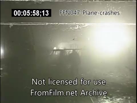 The still depicts a scene from a newsreel related to plane crashes. Visibility appears low due to smoke or fog, with bright lights illuminating the area. Silhouetted figures are seen amidst debris, suggesting the aftermath of an accident. The image conveys a somber and chaotic atmosphere typical of disaster coverage.