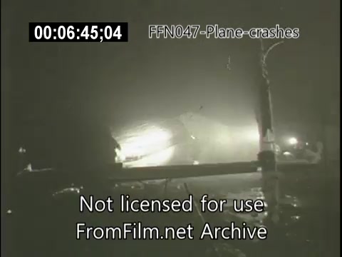 The still from 'FFN047-plane-crashes-newsreel-etc' depicts a scene likely following a plane crash. The image is dimly lit, suggesting either nighttime or low visibility conditions. Debris from the crash is visible, along with emergency lighting, indicating a response to the incident. The atmosphere is somber, highlighting the seriousness of the event.