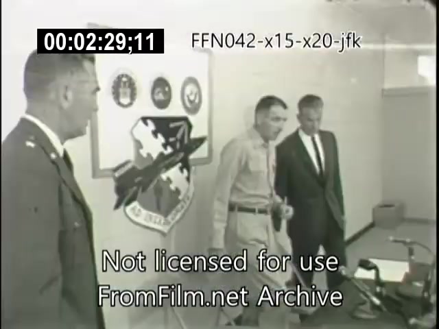 The still depicts a meeting involving military personnel and officials related to the X-15 and X-20 Dynasoar programs. In a formal setting, two men are engaged in discussion, while another stands to the side, possibly presenting information or briefing. There are insignias on the wall that represent the associated military branches or agencies. The atmosphere suggests a focus on aerospace advancements and collaboration.