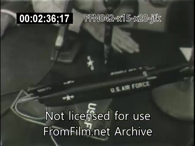 The still features a tabletop setting with a model or object labeled 'U.S. AIR FORCE.' Various microphones are positioned nearby, suggesting a formal presentation or discussion. The setting likely relates to aerospace topics, such as the X-15 and X-20 Dynasoar programs, indicative of the era's space exploration efforts. The visible timecode indicates a specific moment in the footage from the archive.