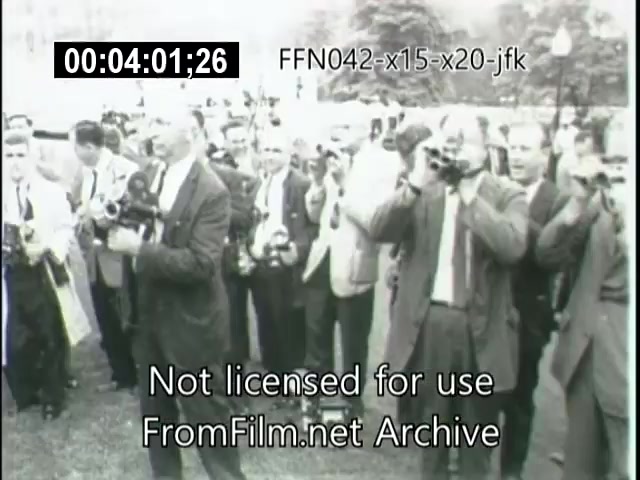 The still captures a group of photographers and journalists, all focused on a singular event, likely involving a significant figure such as JFK. They are equipped with cameras, creating an atmosphere of anticipation and media activity. The setting appears to be outdoors, with trees in the background, signifying an important moment in history possibly related to the X-15 or X-20 Dynasoar aerospace programs. The image is in black and white, emphasizing the archival nature of the footage.