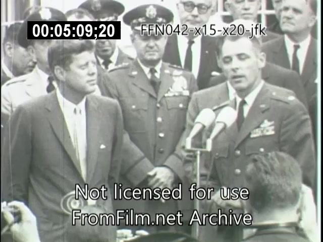 The still captures a moment from a historical event featuring John F. Kennedy, surrounded by military officials. Kennedy appears engaged in conversation with another man in uniform, likely discussing aviation or space program achievements related to the X-15 and X-20 Dynasoar. The scene reflects the significance of aerospace advancements during the early 1960s.