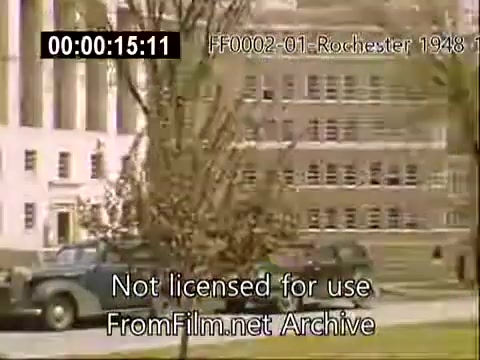 The still features a view of the University of Rochester's campus, likely from the late 1940s to early 1950s. In the foreground, vintage cars are parked along a tree-lined pathway. The architecture of the buildings showcases a blend of mid-century design, with notable structures like the school of medicine visible in the background. The scene captures a tranquil moment in the campus setting during this period, emphasizing the scholarly atmosphere of the university.