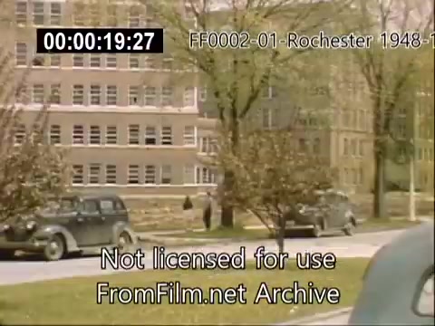 The still features a scene from the University of Rochester, capturing a view of the architecture around the campus during the late 1940s. Several vintage cars are parked along the road, and individuals can be seen walking nearby. The trees are lush, suggesting spring or summer, and the overall ambiance reflects a vibrant university environment of that period. The building depicted likely relates to the Rochester School of Medicine.