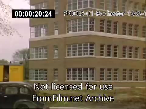 The still from the archival footage depicts a brick building at the University of Rochester, likely part of the School of Medicine, during the late 1940s. The structure has large windows that reflect the era's architectural style. In the foreground, a vintage car is parked, contributing to the historical context of the film. Overall, the image captures a moment in the campus's development during the specified time period.