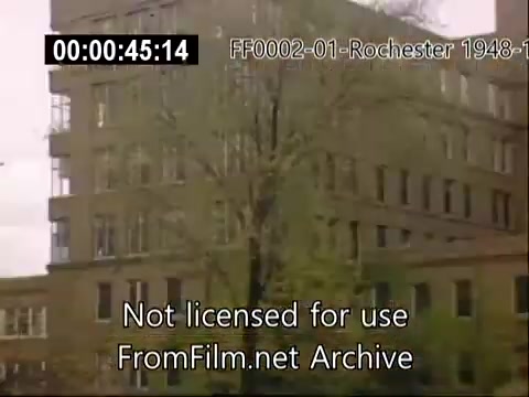 The still captures the exterior of a building associated with the University of Rochester's School of Medicine, set in the late 1940s to early 1950s. The architecture features a mid-century design, with visible windows and a tree in the foreground. The frame conveys a sense of historical significance, highlighting campus life during that era.