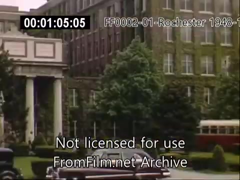 The still shows a scene from the University of Rochester, likely depicting the School of Medicine. The architecture features a prominent building with a classic facade, flanked by well-maintained greenery and trees. A vintage car is parked nearby, and a trolley can be seen in the background, giving it a mid-20th-century ambiance. The overall atmosphere reflects the historical and academic significance of the university during the late 1940s.