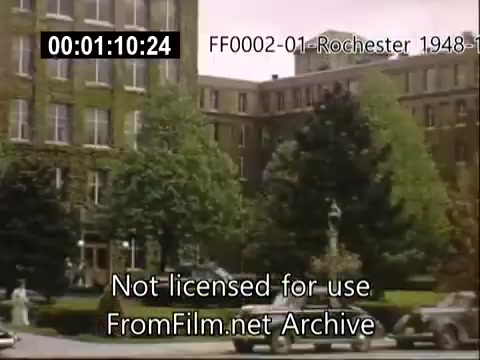 The still depicts a scene from the University of Rochester in the late 1940s. It showcases the architecture of the Rochester School of Medicine, surrounded by greenery and trees. In the foreground, a fountain is visible, along with a couple of parked cars typical of that era. The overall atmosphere reflects a serene campus setting, highlighting the educational environment during that period.