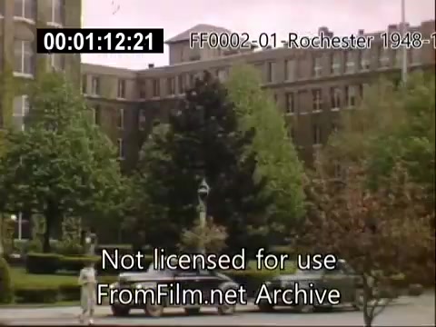 The still features a view of the University of Rochester's School of Medicine from archival footage dated between 1948 and 1950. The scene captures the building's architecture surrounded by lush greenery and trees, with a few people walking in the vicinity. A vintage car is parked in the foreground, indicating the time period. The colors, enhanced by Kodachrome film, provide a vivid representation of campus life during that era.
