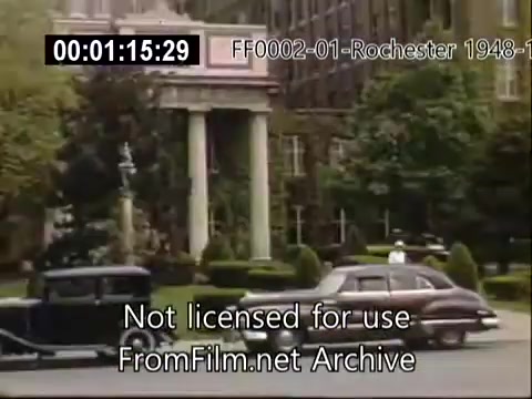 The 16mm still captures a scene from the University of Rochester's campus between 1948 and 1950. It features classic cars parked in front of a stately building with columns, indicative of the era's architecture. Lush greenery and landscaped gardens surround the area, creating a vibrant and welcoming academic environment. The image conveys a sense of mid-20th century campus life, reflecting the historical context of the university during that time.
