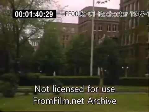 The still depicts the University of Rochester's campus, focusing on the Rochester School of Medicine. It shows green lawns and trees, framed by historical buildings characteristic of mid-20th century architecture. The vibrant colors typical of Kodachrome film capture the serene atmosphere of the university during the late 1940s to early 1950s, reflecting a lively educational environment. Subtitles at the bottom indicate the footage’s archival nature.