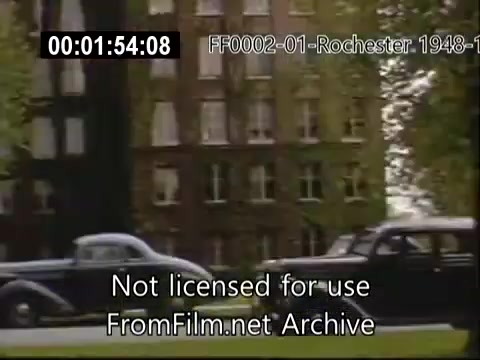The still features vintage automobiles driving past a large, likely historic building at the University of Rochester, captured in rich Kodachrome color typical of the late 1940s to early 1950s. The architecture suggests a classical style, characteristic of academic institutions, surrounded by greenery. The scene reflects everyday life on campus during that era, showcasing the environment around the Rochester School of Medicine.