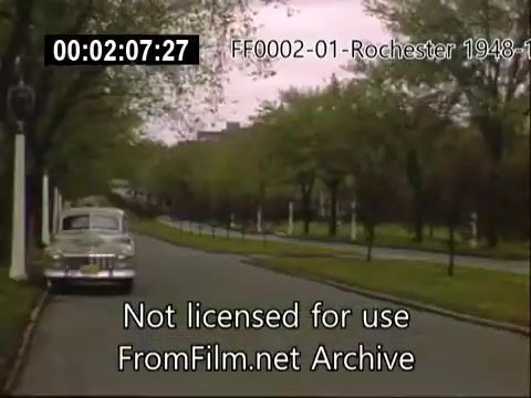 The 16mm still from 'University of Rochester (NY) archival footage - 1948-1950 Kodachrome' depicts a scene along a tree-lined street, likely on or near the University of Rochester campus. A classic car drives along the road, illustrating the mid-20th century setting. The greenery and pleasant atmosphere hint at a peaceful day, capturing a snapshot of campus life during that era. The footage is part of home movies taken in and around the university during the late 1940s to early 1950s.