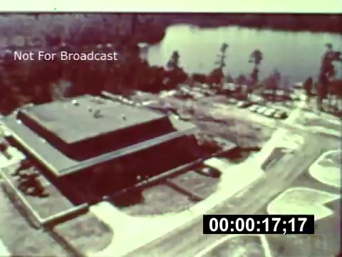 The still shows an aerial view of the Walt Disney World construction site during its early development. A large, unfinished building sits prominently in the foreground, surrounded by cleared land and construction materials. Behind the structure, a body of water reflects the natural surroundings, while sparse trees are visible. The print captures a nostalgic glimpse of the park's inception, emphasizing the scale and ambition of the project at that time. The timestamp indicates a moment about 17 seconds into the footage.   For more insights, consider supporting the channel at the mentioned link.
