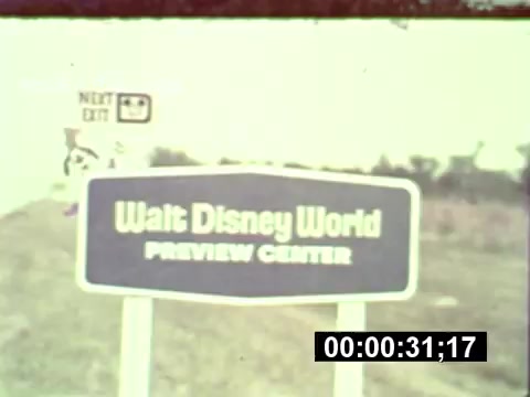 The still from the 16mm print captures a sign for the 'Walt Disney World Preview Center.' The sign is prominently displayed against a backdrop of undeveloped land, indicative of the park's construction phase. The color and quality reflect the vintage nature of the film, typical of the era. The sign shares its purpose clearly, as a glimpse into the upcoming major attraction. The frame also includes a timestamp, emphasizing its role in documenting this historic moment.