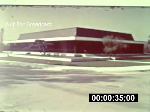 The still from the 16mm print of 'WDW preview' shows a modern, angular building set against a clear sky. The structure has a flat roof and is surrounded by minimal landscaping, suggesting it was a new development. The frame includes a timestamp indicator, reinforcing its historical context as part of the construction phase of Walt Disney World. Overall, the image captures a moment in the early stages of the park's establishment.