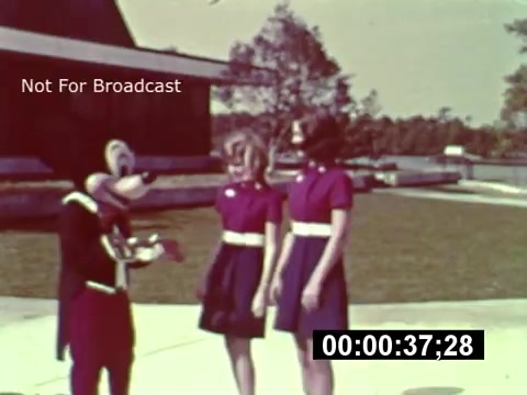 The still from the 16mm print of 'WDW Preview' shows an early promotional moment for Walt Disney World, capturing a costumed character, likely from Disney, interacting with two young women in matching outfits. The background hints at the park's construction with a clear sky and trees. The image is warm and nostalgic, showcasing the excitement and anticipation surrounding the park's opening. The timecode suggests it's part of a larger film segment.