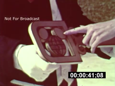 The still from the 16mm print of 'WDW Preview' shows a person holding a framed design or logo, likely related to the Walt Disney World project during its construction phase. The individual is pointing to a globe icon within the design, emphasizing its significance. The image has a vintage aesthetic, typical of the era, and features a 'Not For Broadcast' watermark, along with a timecode at the bottom. This snapshot captures a moment of excitement and anticipation as the iconic theme park was being brought to life.