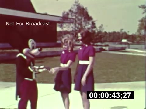 The still from the 16mm print features a character in a costume, likely a Disney character, interacting with two women dressed in matching purple outfits with white belts. The setting appears to be outdoors, with a grassy area and some background trees, hinting at a construction phase of Walt Disney World. The overall vibe suggests a promotional or preview event during the park's development. The watermark indicates it's not for broadcast use, and a timestamp at the bottom suggests it's part of a longer film sequence.