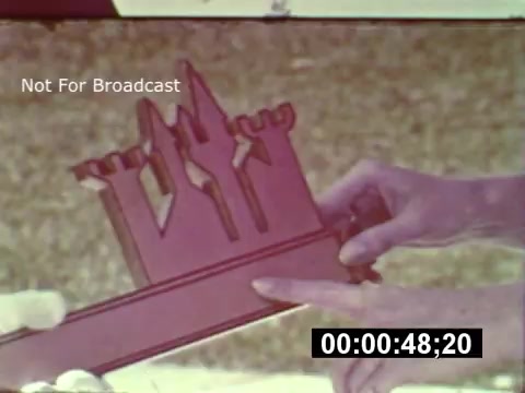 The 16mm still showcases a close-up of a hand presenting a model of a castle structure, likely part of a preview related to the construction of Walt Disney World. The model features distinctive turret-like features, indicating it may represent a themed attraction within the park. The background is blurred, with a green lawn suggesting an outdoor setting, potentially during a promotional event or presentation. The frame includes a timestamp and a watermark indicating it is not for broadcast.