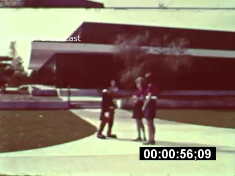 The still from the 16mm print of 'WDW Preview' captures a moment during the construction phase of Walt Disney World. In the foreground, a small group of individuals stands on a walkway, engaged in conversation, while a modern building looms in the background, reflective of the architectural style of the time. The vibrant colors and composition highlight the excitement and anticipation surrounding the park's development. The scene conveys a sense of nostalgia and curiosity about the future of the park.