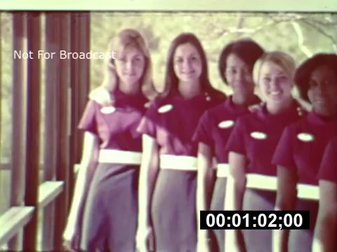 The still depicts a group of young women dressed in matching uniforms, standing together in a bright, welcoming environment. They all appear to be smiling directly at the camera, showcasing a sense of camaraderie and excitement. The backdrop suggests a construction setting typical of the early stages of Walt Disney World's development, hinting at the park's vibrant future. The footage is marked with 'Not For Broadcast' and features a timestamp, indicating it's an archival piece from the project’s promotion.