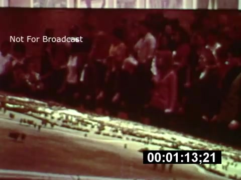 The still captures a moment from a 16mm preview of Walt Disney World during its construction phase. In the foreground, a crowd is visible, likely watching a presentation or display about the park. Behind them, a detailed model or aerial view of the park's layout is shown, illustrating the planned development. The image highlights the excitement and anticipation surrounding the park's upcoming opening, capturing a nostalgic moment in Disney history.