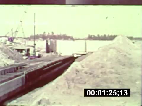 The still from the 16mm print 'WDW preview - from when the park was under construction' shows a construction site for Walt Disney World. In the foreground, large mounds of sandy material are visible, indicative of excavation work. There are outlines of structures being built, along with construction equipment in the background. This image captures a moment in the early development phase of the theme park, highlighting the scale of the construction efforts. The color tone appears slightly faded, characteristic of older film prints.