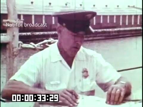 The still from 'Immigrant Heritage' features a man in a white uniform, likely a customs or immigration officer, seated at a table near a body of water. He appears to be reviewing documents, possibly related to immigration processes. The background includes a shipping dock, suggesting a connection to immigration or arrival by sea. Screen text indicates the clip is not for broadcast, with a timecode visible at the bottom. The film aims to educate about immigrant experiences, potentially highlighting historical contexts involving Irish immigrants.